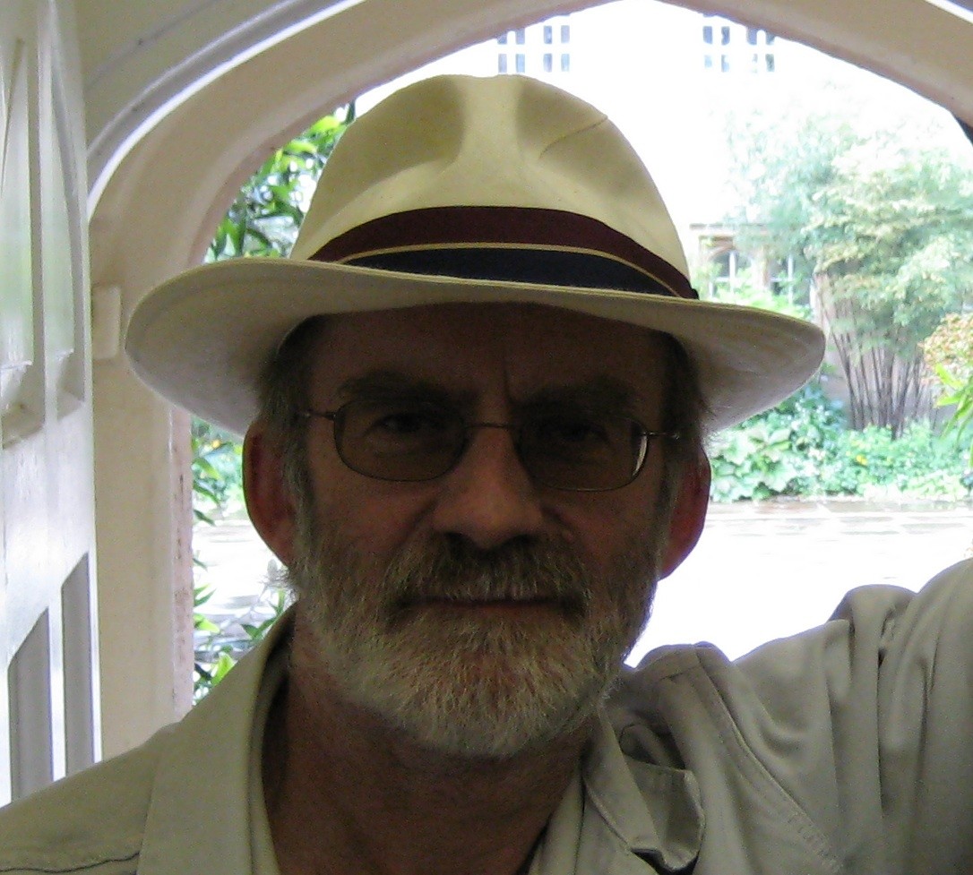 A white man with a grey beard and glasses looks at the camera. He wears a white hat with a red band.