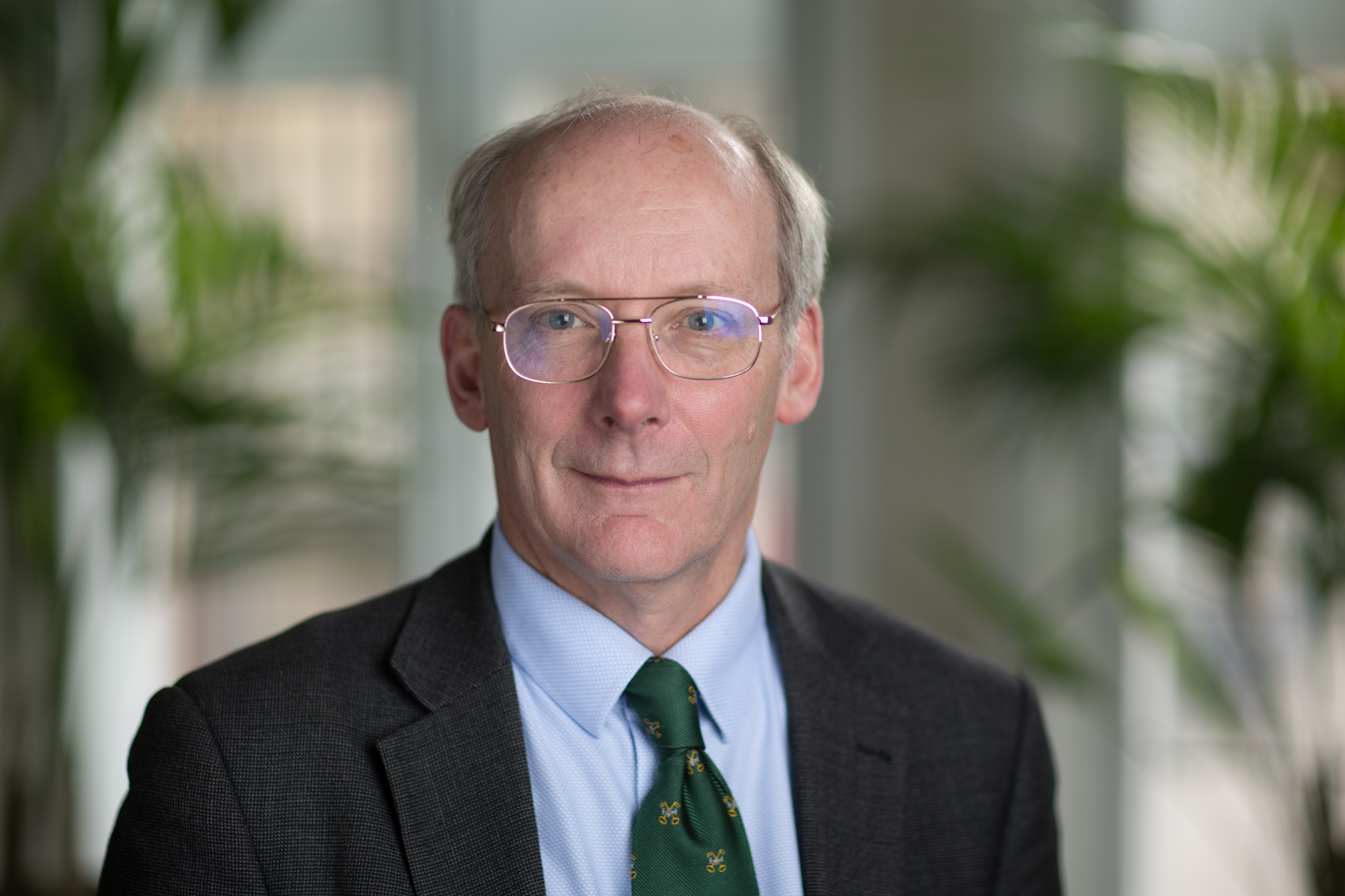 A white man wearing glasses smiles at the camera. He is dressed in a suit with a green tie.