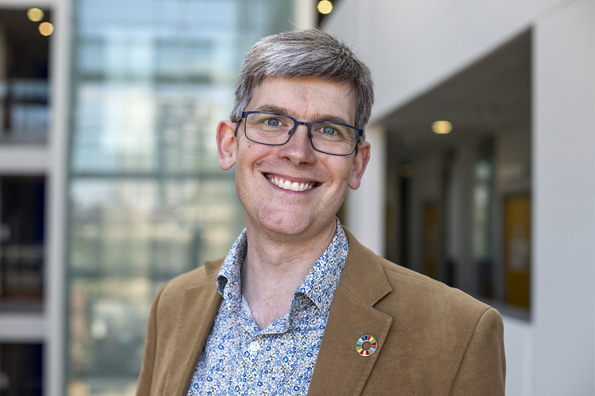 A white man with grey hair and glasses smiles at the camera. He wears a khaki coloured blazer and patterned blue shirt