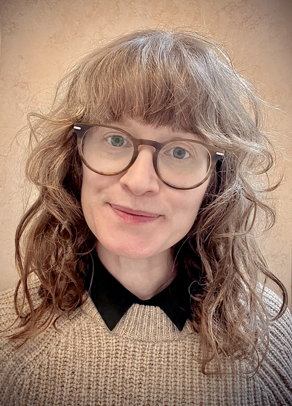 A white woman with dark curly hair and blue eyes smiles toward the camera. She is wearing glasses and a beige fisherman jersey.