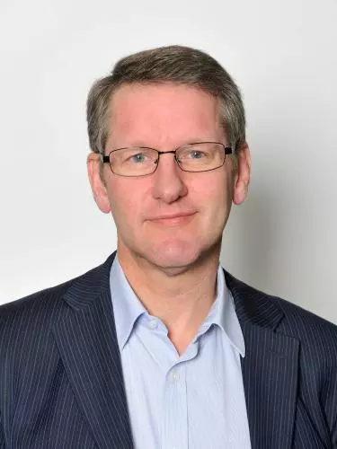 A white man with blonde hair and blue eyes smiles at the camera. He wears glasses and a blue shirt and striped blazer.