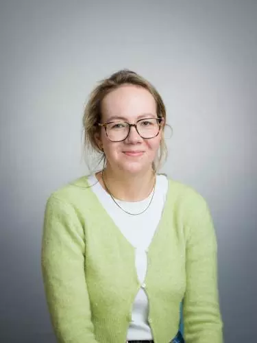A white woman with blonde hair smiles at the camera. She is wearing a yellow cardigan and glasses.