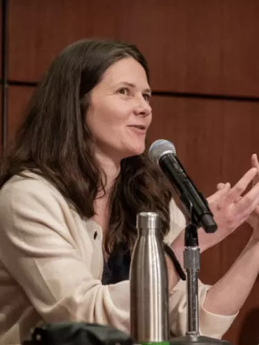 A white woman with dark hair and wearing a cream cardigan, talks with her hands. She has a microphone in front of her.