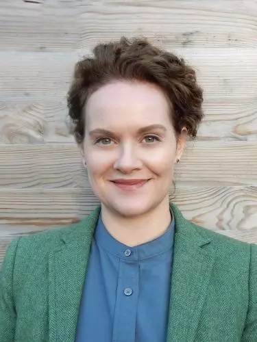 A dark-haired white woman smiles against a neutral backdrop. She is wearing a green jacket and blue shirt.