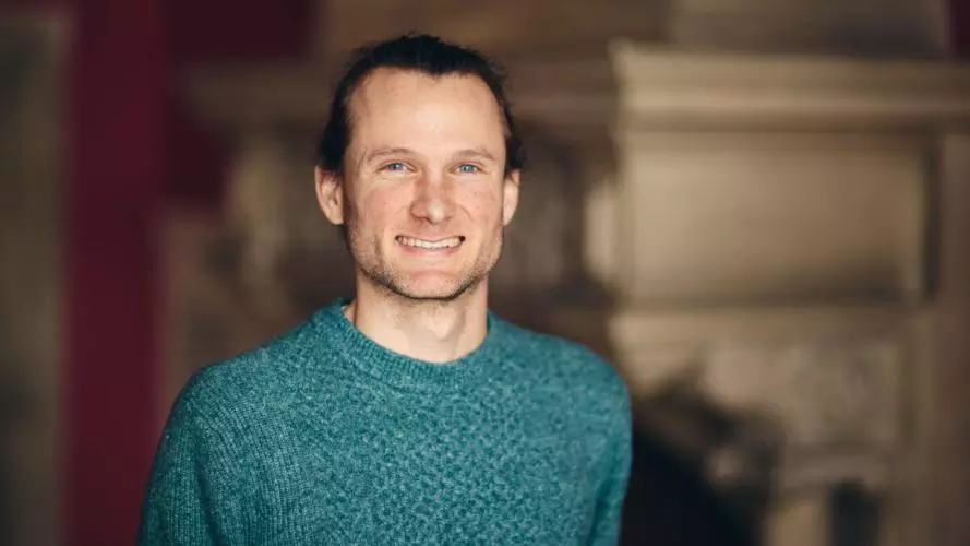 A white man with dark hair tied back smiles at the camera. He wears a seagreen jumper.