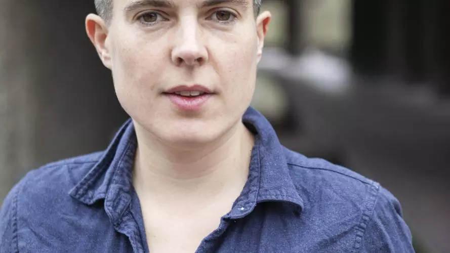 A white woman smiles looking at the camera. She has short black hair and is wearing a dark blue top with a colour and buttons.