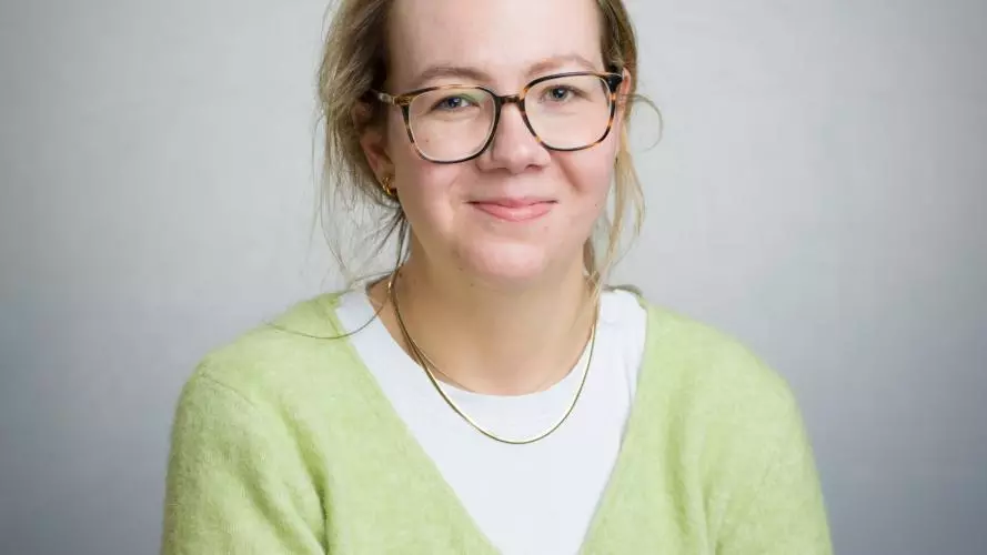 A white woman with blonde hair smiles at the camera. She is wearing a yellow cardigan and glasses.