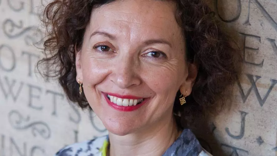 A white woman with brown curly hair smiles at the camera. She wears a blue and yellow-green shirt.