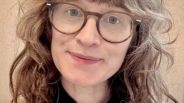 A white woman with dark curly hair and blue eyes smiles toward the camera. She is wearing glasses and a beige fisherman jersey.