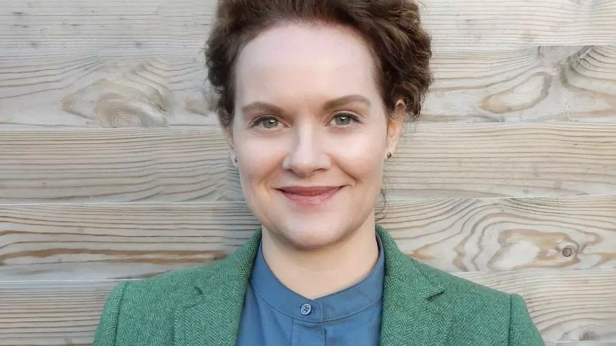 A dark-haired white woman smiles against a neutral backdrop. She is wearing a green jacket and blue shirt.