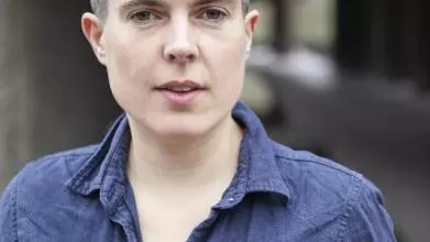 A white woman smiles looking at the camera. She has short black hair and is wearing a dark blue top with a colour and buttons.