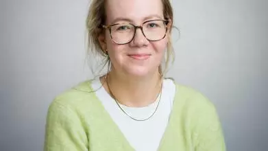 A white woman with blonde hair smiles at the camera. She is wearing a yellow cardigan and glasses.