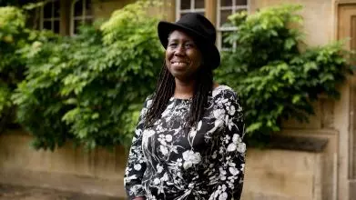 A black woman with long braids smiles at the camera. She wears a black hat and a black and white patterned dress.