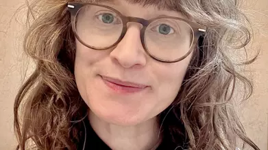 A white woman with dark curly hair and blue eyes smiles toward the camera. She is wearing glasses and a beige fisherman jersey.