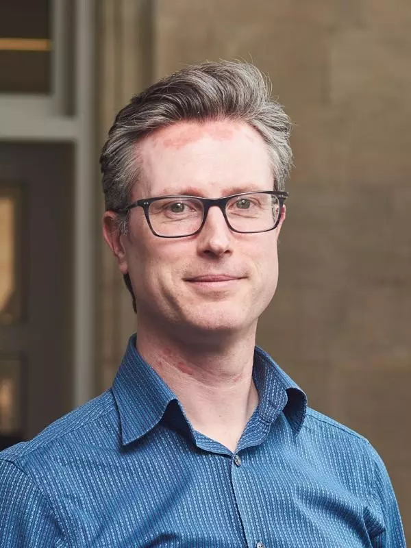A white man with glasses smiles at the camera. He has greying hair and wears a blue shirt