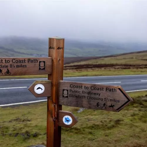 A sign showing the directions for the Wainwright Way, including the distinctive 'AW' initials at the top of the sign. Behind the sign is road and then rolling hills shrouded in mist.