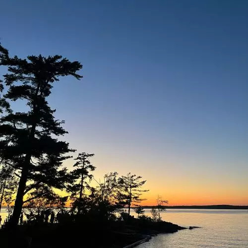The sun sets over a lake. In darkness in the left corner are trees and the lake edge.