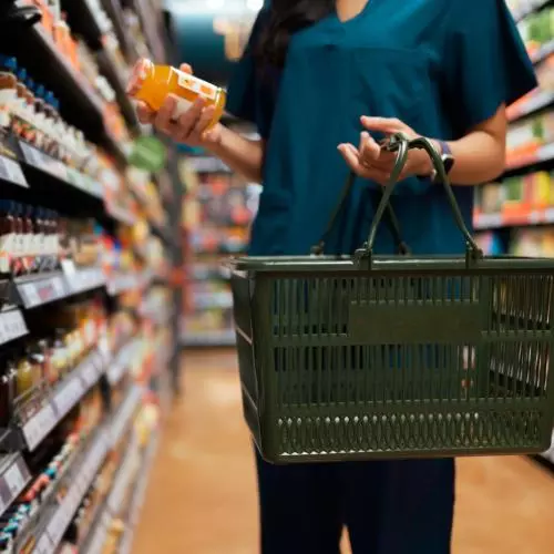 A person in a dark green shirt holds a green shopping basket. The basket is empty. In their right hand they hold a jar of something yellow.