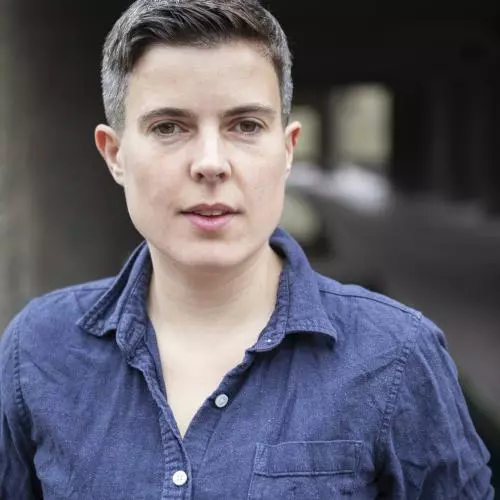 A white woman smiles looking at the camera. She has short black hair and is wearing a dark blue top with a colour and buttons.