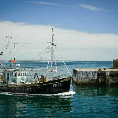 A small boat enters a walled harbour. The sea is blue with some white cloud on the horizon. A lighthouse stands on the harbour wall.