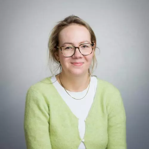 A white woman with blonde hair smiles at the camera. She is wearing a yellow cardigan and glasses.