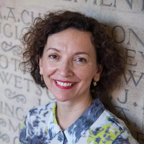 A white woman with brown curly hair smiles at the camera. She wears a blue and yellow-green shirt.