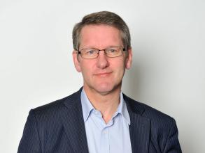 A white man with blonde hair and blue eyes smiles at the camera. He wears glasses and a blue shirt and striped blazer.