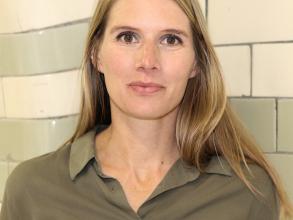 A white woman with long blonde hair smiles at the camera. She is wearing a brown shirt.