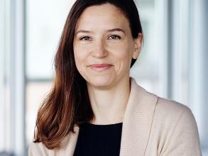 A white woman with long dark hair smiles at the camera. She wears a cream cardigan over a black shirt.